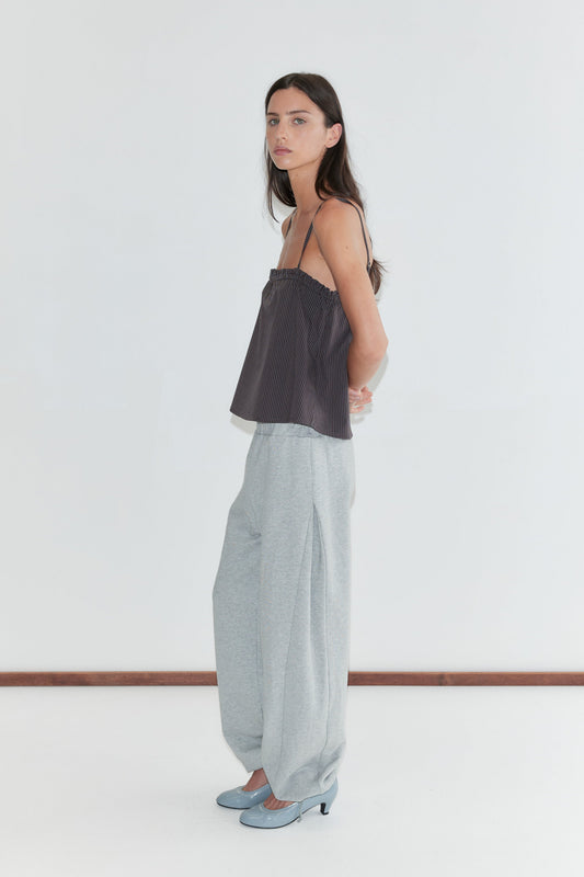 A woman with long dark hair stands in profile against a plain white background wearing the Frill Placket Top - Brown Blue Stripe by Deiji Studios, a dark brown fine-striped cami top, with grey marle trousers and pale blue heels.