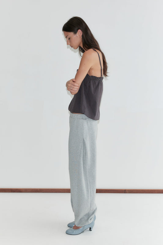 A woman with long dark hair stands in profile against a plain white background, looking down, wearing the Frill Placket Top - Brown Blue Stripe by Deiji Studios, a dark brown fine-striped cami top, with grey marle trousers and pale blue heels.