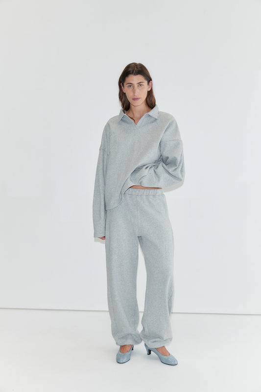 A person stands against a white background wearing the Jersey Jumper - Grey Marle, styled with matching sweatpants and blue heels, with one hand tucked into their pocket.