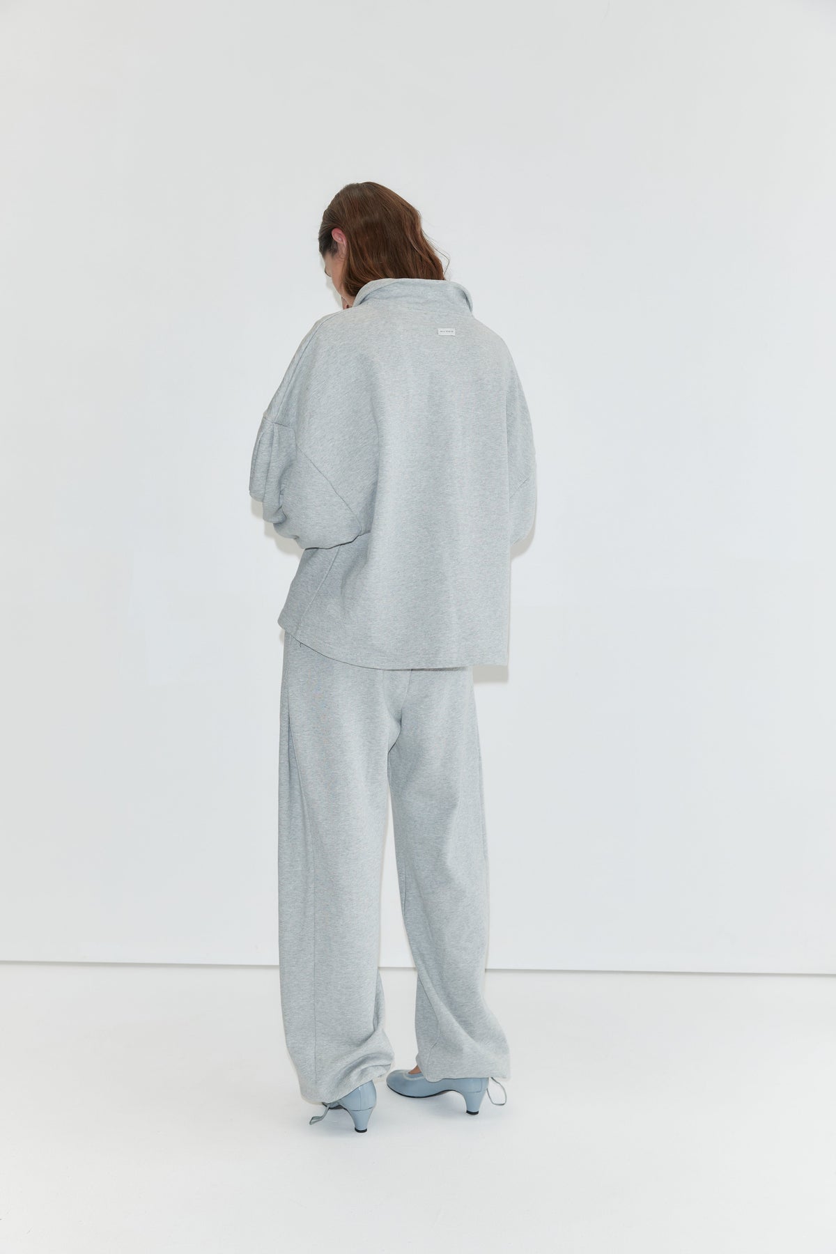A person stands facing a white wall, wearing the Jersey Jumper - Grey Marle, a loose light gray cotton jersey jumper with an open polo collar, paired with matching pants and light blue heeled shoes. Their long hair partially covers their face.