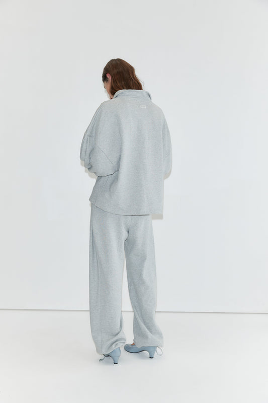 A person stands facing a white wall, wearing the Jersey Jumper - Grey Marle, a loose light gray cotton jersey jumper with an open polo collar, paired with matching pants and light blue heeled shoes. Their long hair partially covers their face.