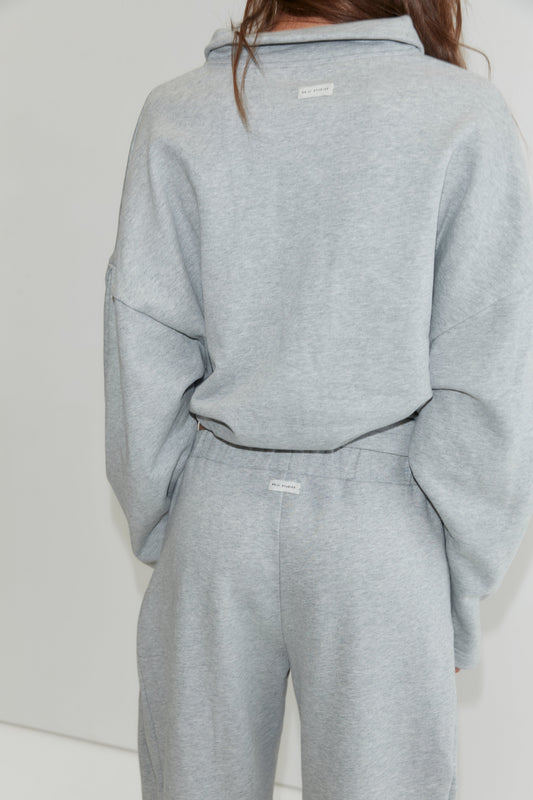 A person wearing a matching light gray sweatshirt and wide-leg cotton pant set, shown from the back. The outfit, inspired by Deiji Studios, features white tags on the upper back of the top and on the waistband of the Side Pleat Pant - Grey Marle.