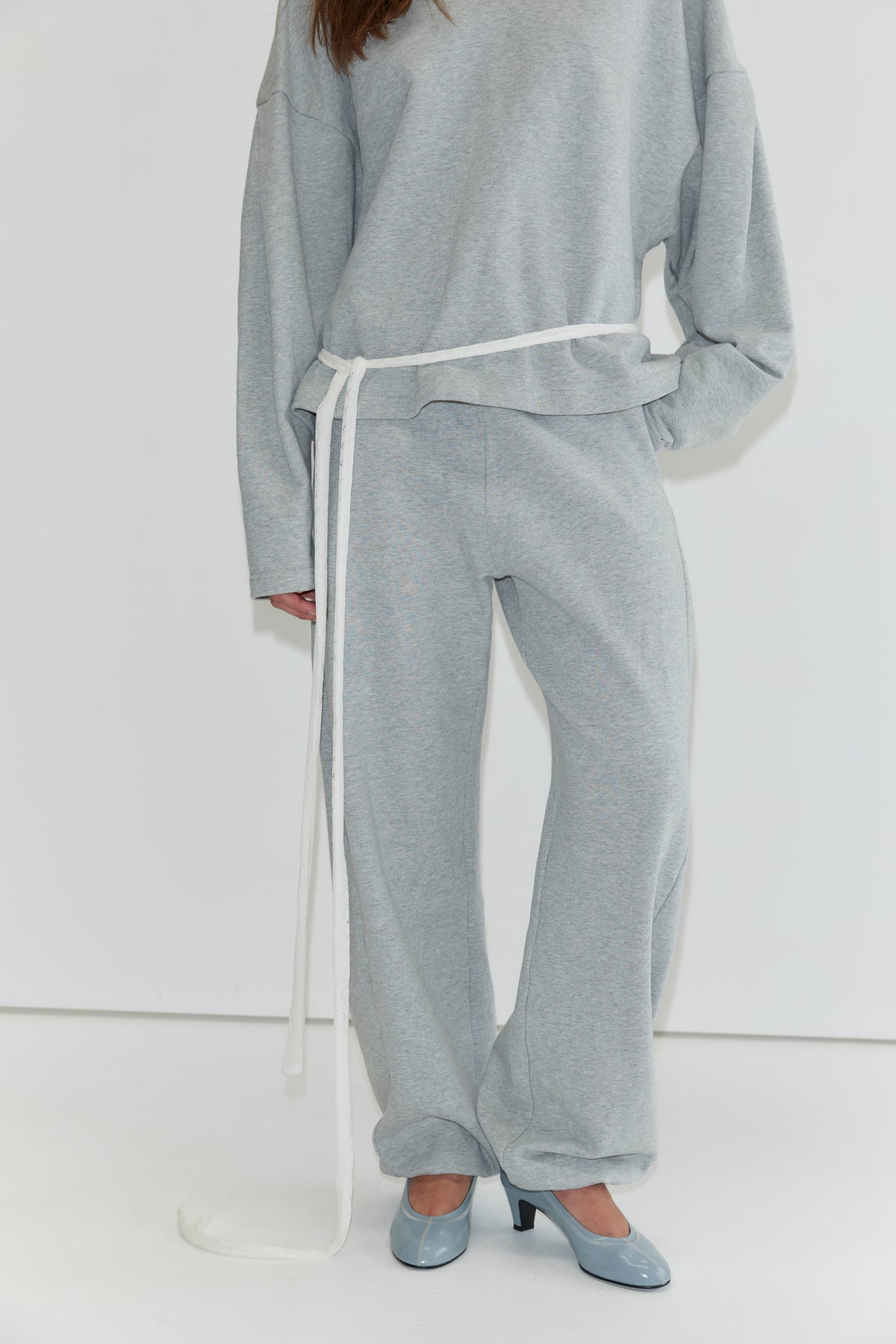 A person is shown wearing the Side Pleat Pant - Grey Marle, styled with a loose-fitting light gray sweatshirt and cinched at the waist with a long white belt, paired with blue high-heeled shoes. The face is not visible, capturing the relaxed elegance characteristic of Deiji Studios collections.