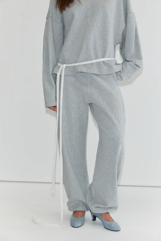 A person is shown wearing the Side Pleat Pant - Grey Marle, styled with a loose-fitting light gray sweatshirt and cinched at the waist with a long white belt, paired with blue high-heeled shoes. The face is not visible, capturing the relaxed elegance characteristic of Deiji Studios collections.