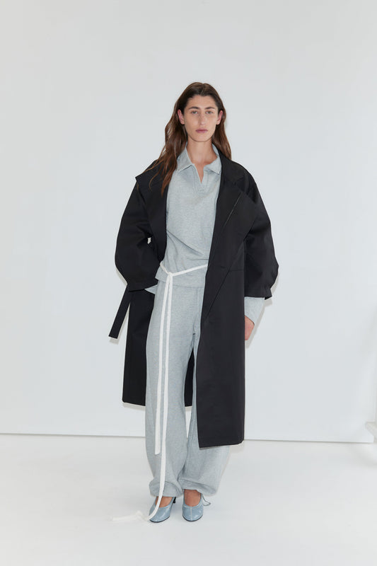 Wearing the Jersey Jumper in Grey Marle against a white background, the person completes their look with an open polo collar design, a white belt, a long black coat, and light blue shoes.