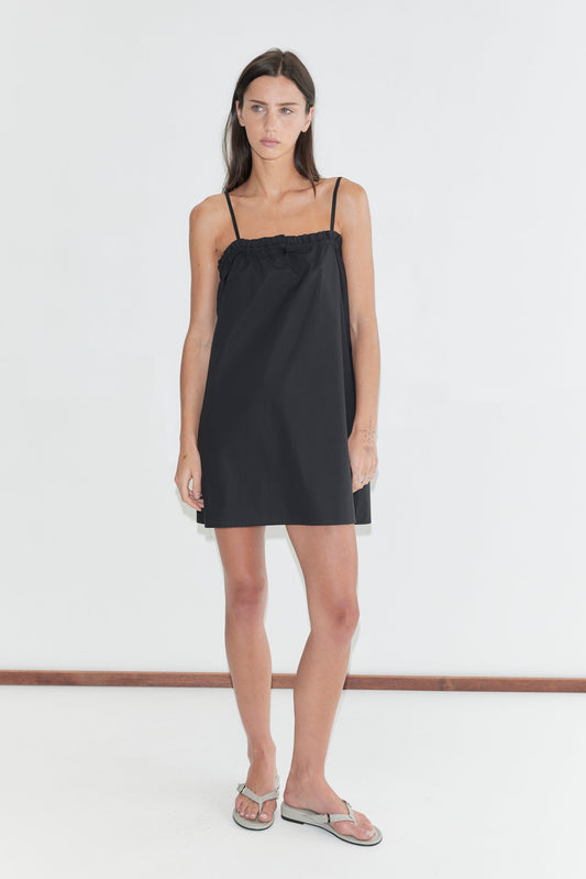 A woman with long dark hair stands against a plain white background wearing the Frill Placket Dress - Black by Deiji Studios, a black A-line mini dress with thin straps and a frilled placket neckline, with grey flip-flops.