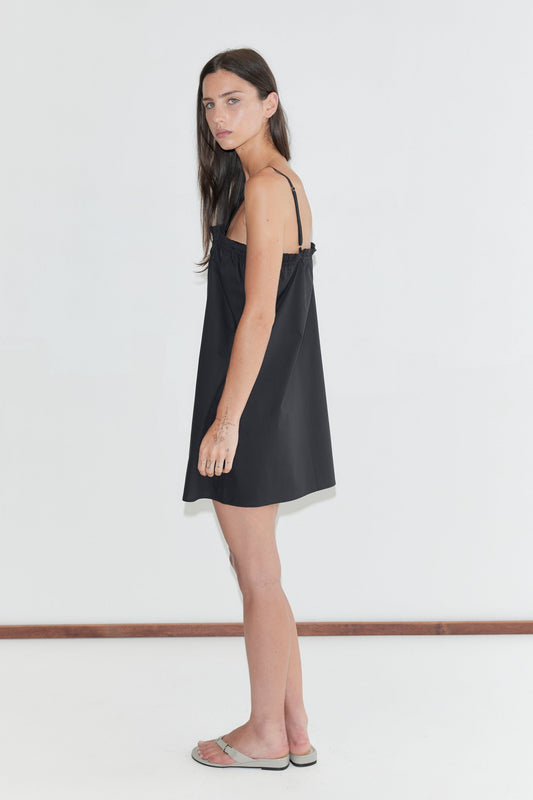 A woman with long dark hair stands in profile against a plain white background wearing the Frill Placket Dress - Black by Deiji Studios, a black A-line mini dress with thin straps, and flip-flops.