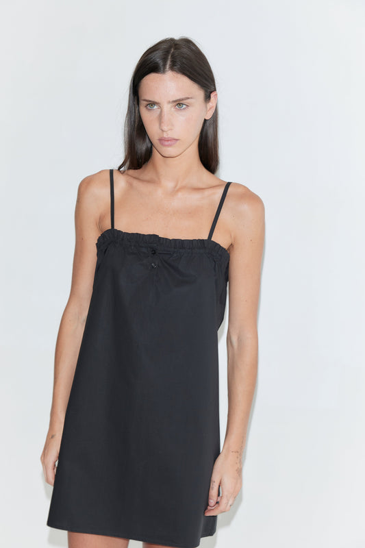 A woman with long dark hair stands against a plain white background wearing the Frill Placket Dress - Black by Deiji Studios, a black mini dress with thin straps and a gathered, buttoned frill placket at the neckline.