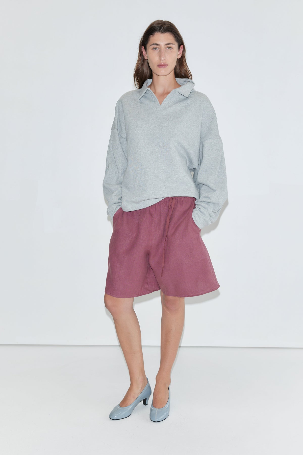 A person wearing a light gray sweatshirt, the "Half Short - Brick" linen shorts with an elasticated waistband and wide leg opening in a knee-length style, and blue heels stands against a plain white background, facing the camera with hands in their pockets.