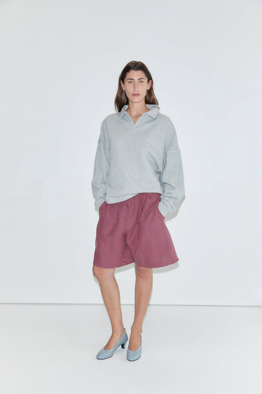 A person wearing a light gray sweatshirt, the "Half Short - Brick" linen shorts with an elasticated waistband and wide leg opening in a knee-length style, and blue heels stands against a plain white background, facing the camera with hands in their pockets.
