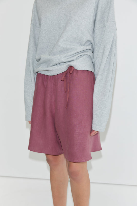 A person wearing a loose grey long-sleeve shirt and high-waisted, knee-length Half Short - Brick relaxed linen shorts with an elasticated waistband and wide leg opening, standing against a plain white background.