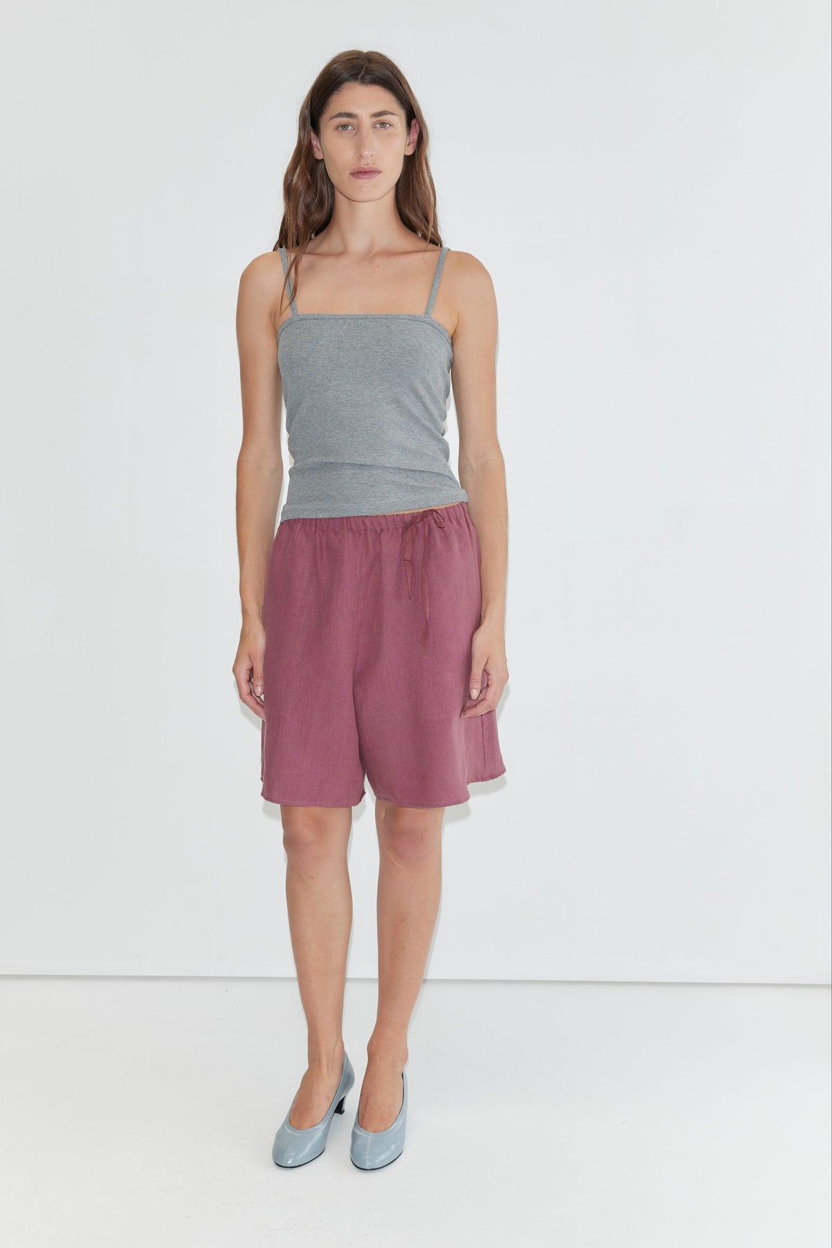 A person stands against a plain white background, wearing a gray spaghetti strap top, Half Short - Brick with an elasticated waistband and wide leg opening in maroon, paired with light blue shoes.