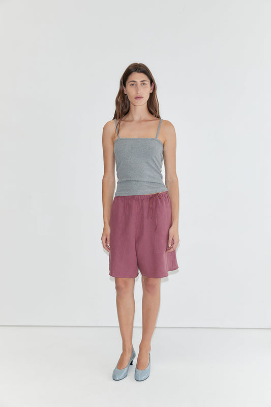 A person stands against a plain white background, wearing a gray spaghetti strap top, Half Short - Brick with an elasticated waistband and wide leg opening in maroon, paired with light blue shoes.