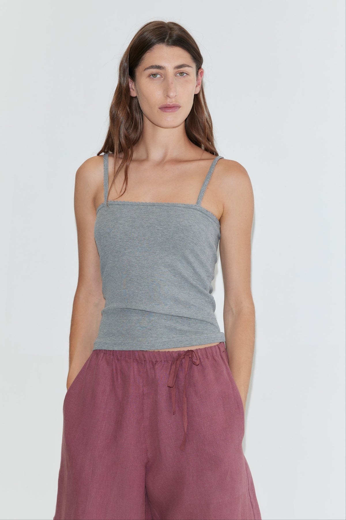A woman with long brown hair, dressed in a gray spaghetti strap top and the Half Short - Brick, which features a wide leg opening and an elasticated waistband, stands against a plain white background with her hands in her pockets.