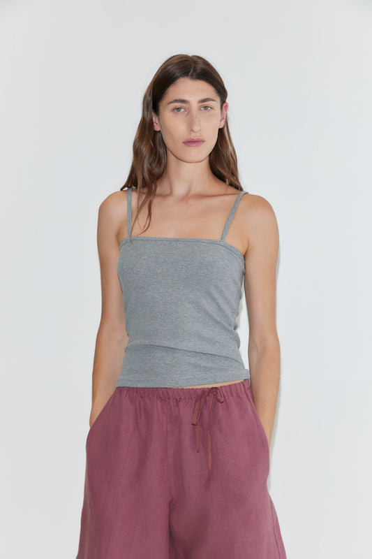 A woman with long brown hair, dressed in a gray spaghetti strap top and the Half Short - Brick, which features a wide leg opening and an elasticated waistband, stands against a plain white background with her hands in her pockets.