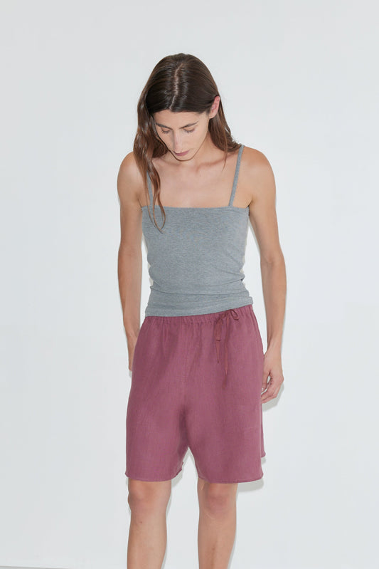 A person with long brown hair, wearing the Scallop Tank - Grey and loose maroon shorts, looks down while standing against a plain white background.