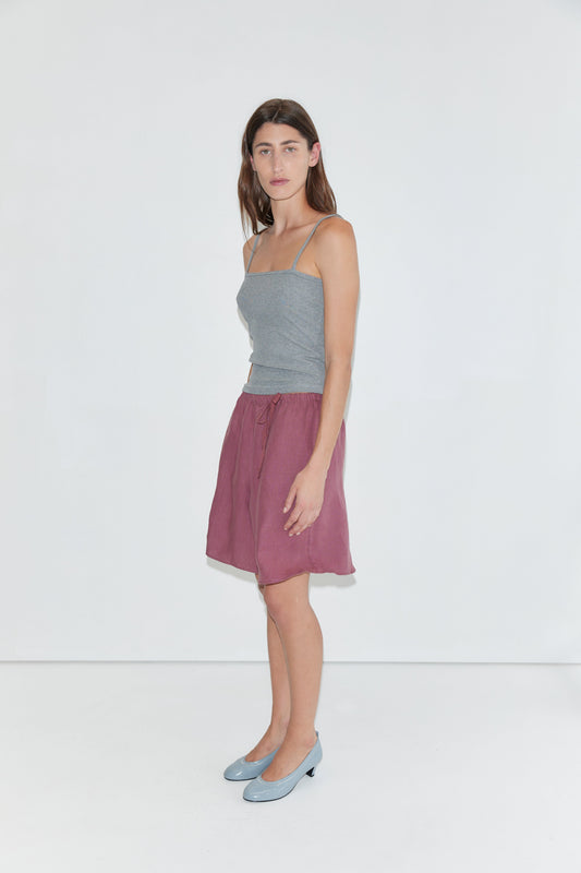A woman stands against a plain white background, wearing the Scallop Tank - Grey with spaghetti straps, paired with a mauve knee-length skirt and light blue low-heeled shoes.
