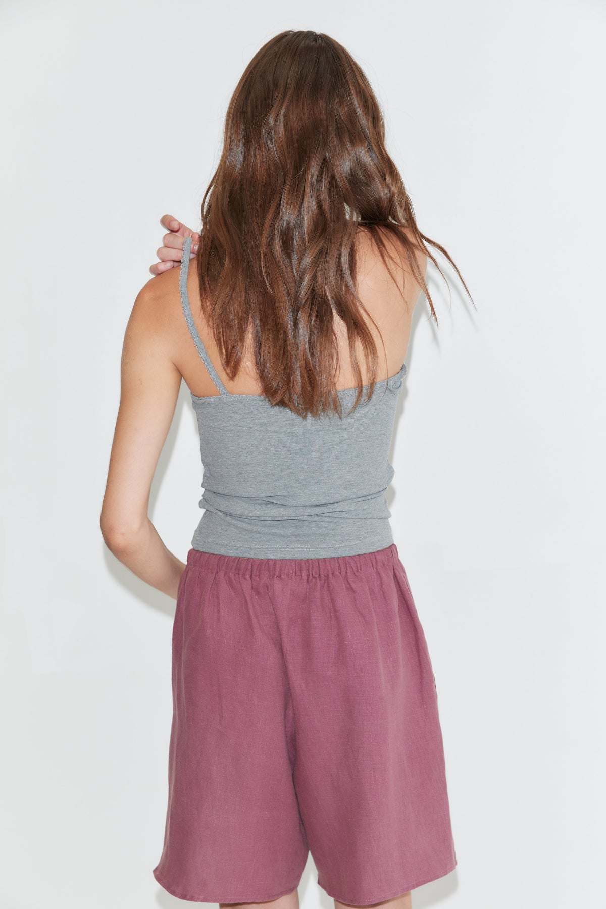 Wearing a gray tank top and the Half Short - Brick, which features an elasticated waistband and wide leg opening, a person with long brown hair stands facing away from the camera against a plain white background.