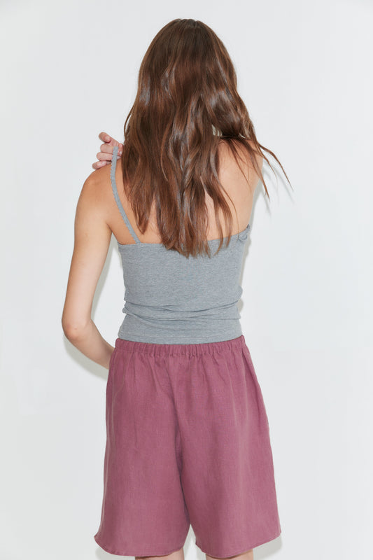 Wearing a gray tank top and the Half Short - Brick, which features an elasticated waistband and wide leg opening, a person with long brown hair stands facing away from the camera against a plain white background.
