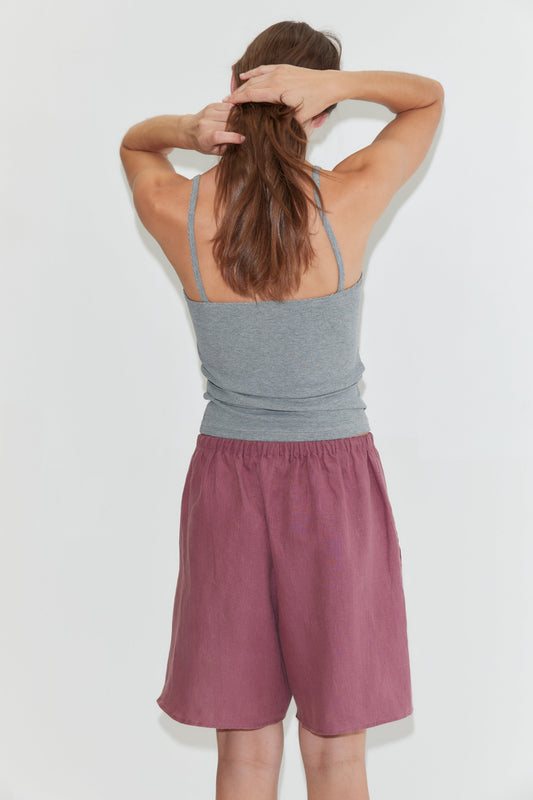 Wearing the Scallop Tank in Grey, a person with long brown hair, paired with mauve shorts, stands facing away and ties their hair with both hands against a plain white background.