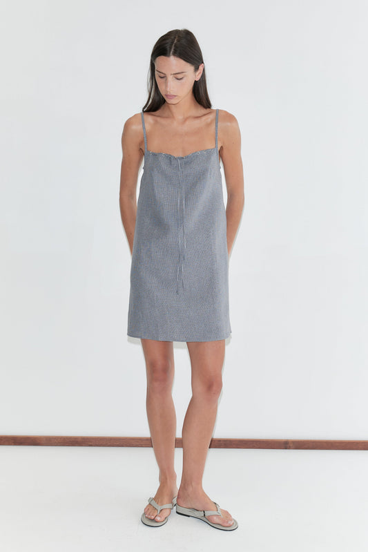 A woman with long dark hair stands against a plain white background looking down, wearing the Weave Mini - Indigo Check by Deiji Studios, an indigo micro-check linen mini dress with thin straps, and flip-flops.
