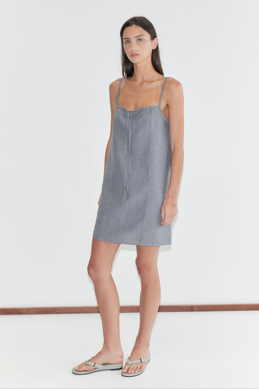 A woman with long dark hair stands against a plain white background wearing the Weave Mini - Indigo Check by Deiji Studios, an indigo micro-check linen mini dress with thin straps, and flip-flops.