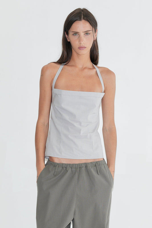 A woman with long dark hair stands against a plain white background wearing the Folded Top - Cloud Stripe by Deiji Studios, a pale grey-blue fine-striped fold-over halter top, with grey check wide-leg trousers and flip-flops.