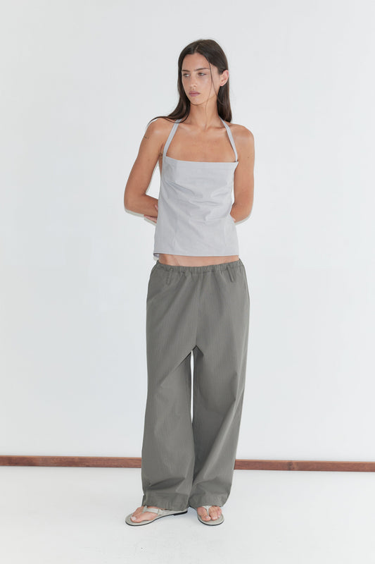 A woman with long dark hair stands against a plain white background wearing the Folded Top - Cloud Stripe by Deiji Studios, a pale grey-blue fine-striped fold-over halter top, with grey check wide-leg trousers and flip-flops.