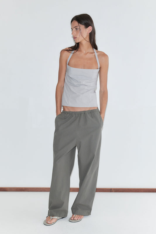 A woman with long dark hair stands against a plain white background wearing the Hem Toggle Pant - Moss Stripe by Deiji Studios, moss-green fine-check trousers, with a pale grey striped halter top.