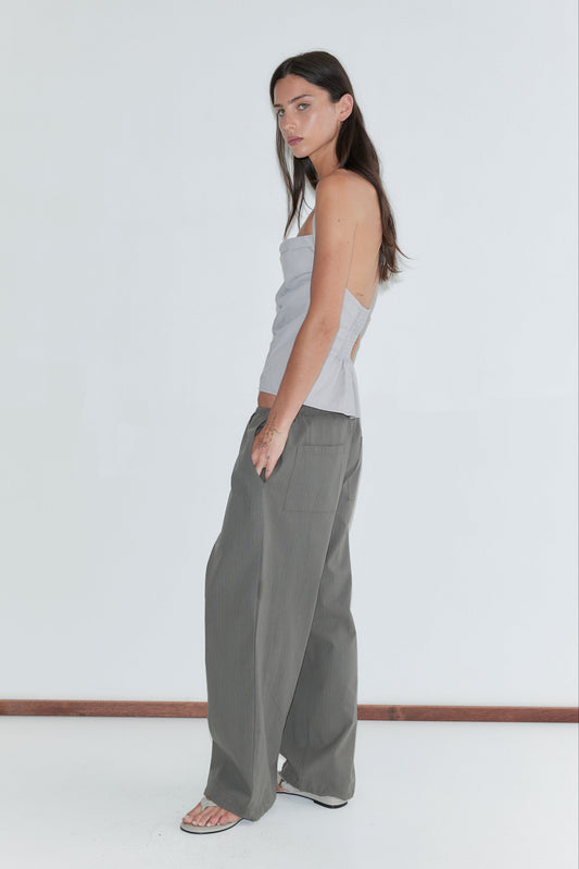 A woman with long dark hair stands in profile against a plain white background wearing the Hem Toggle Pant - Moss Stripe by Deiji Studios, moss-green fine-check trousers with back patch pockets, and a pale grey striped halter top.