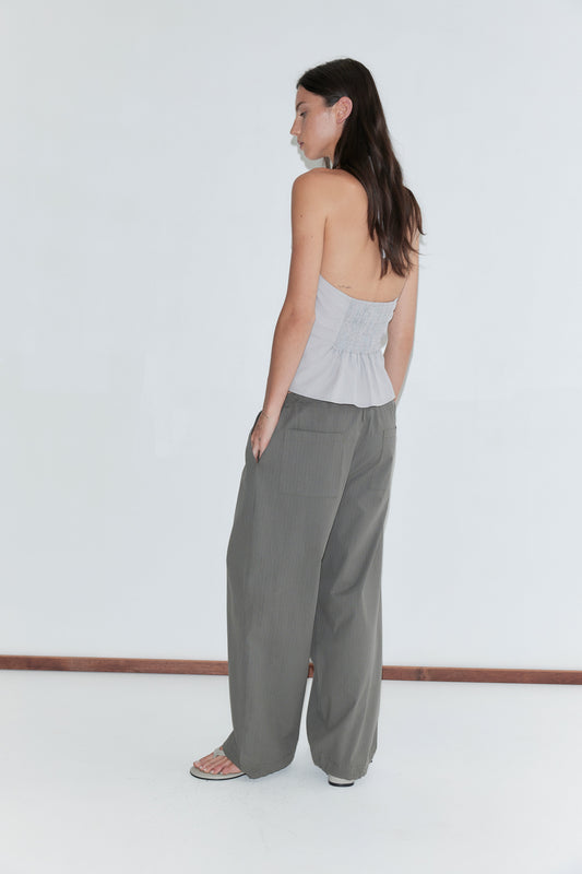 A woman with long dark hair stands with her back to the camera against a plain white background wearing the Folded Top - Cloud Stripe by Deiji Studios, a pale grey-blue fine-striped fold-over halter top, with grey check trousers.