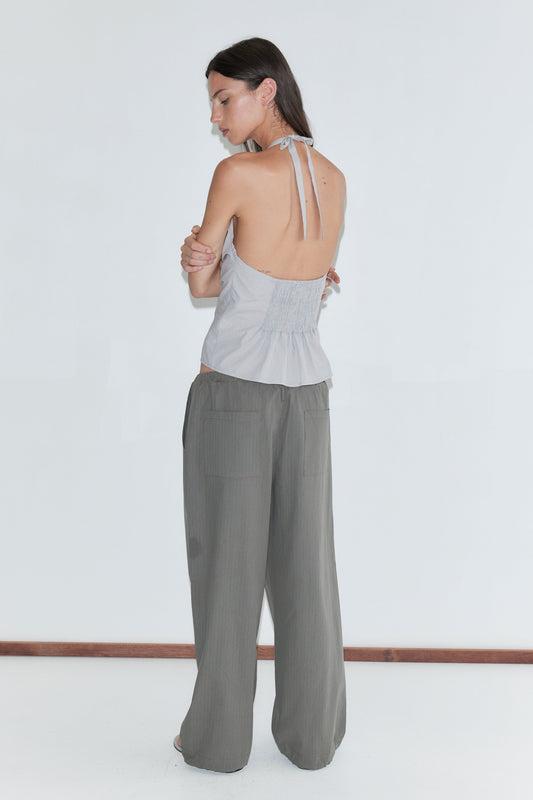 A woman with long dark hair stands with her back to the camera against a plain white background wearing the Hem Toggle Pant - Moss Stripe by Deiji Studios, moss-green fine-check trousers with back patch pockets, and a pale grey striped halter top.