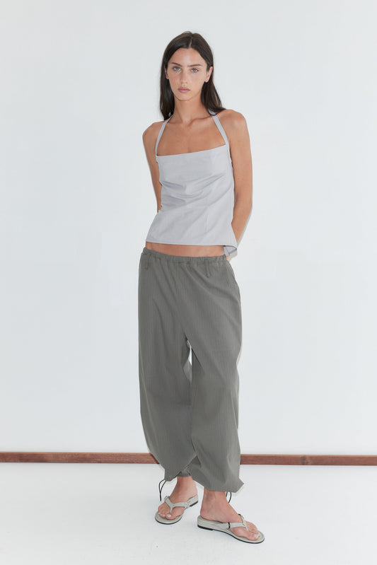 A woman with long dark hair stands against a plain white background wearing the Hem Toggle Pant - Moss Stripe by Deiji Studios, moss-green fine-check trousers with a toggle-gathered hem, with a pale grey striped halter top and flip-flops.