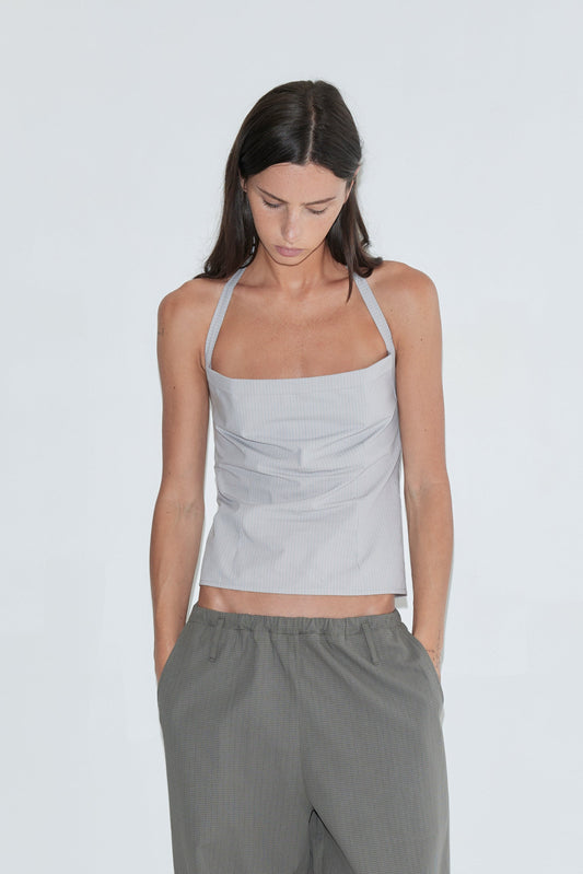 A cropped view of a woman looking down, wearing the Folded Top - Cloud Stripe by Deiji Studios, a pale grey-blue fine-striped fold-over halter top, with grey check trousers against a plain white background.