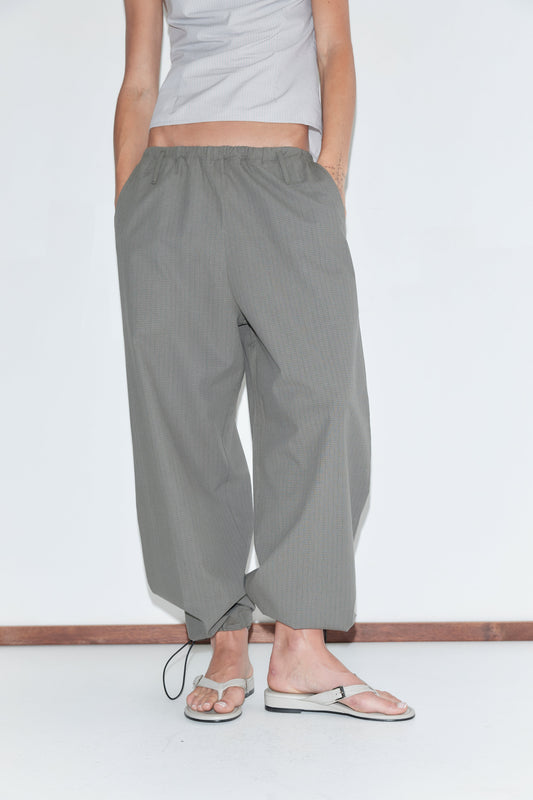 A cropped view of the Hem Toggle Pant - Moss Stripe by Deiji Studios, moss-green fine-check trousers with a toggle-gathered hem, worn with flip-flops against a plain white background.