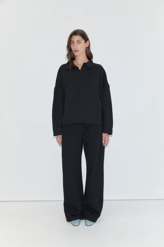 A person with long brown hair is wearing the Jersey Jumper - Black, a loose-fitting cotton piece with an open polo collar, paired with black pants and light-colored shoes, standing against a plain white background.