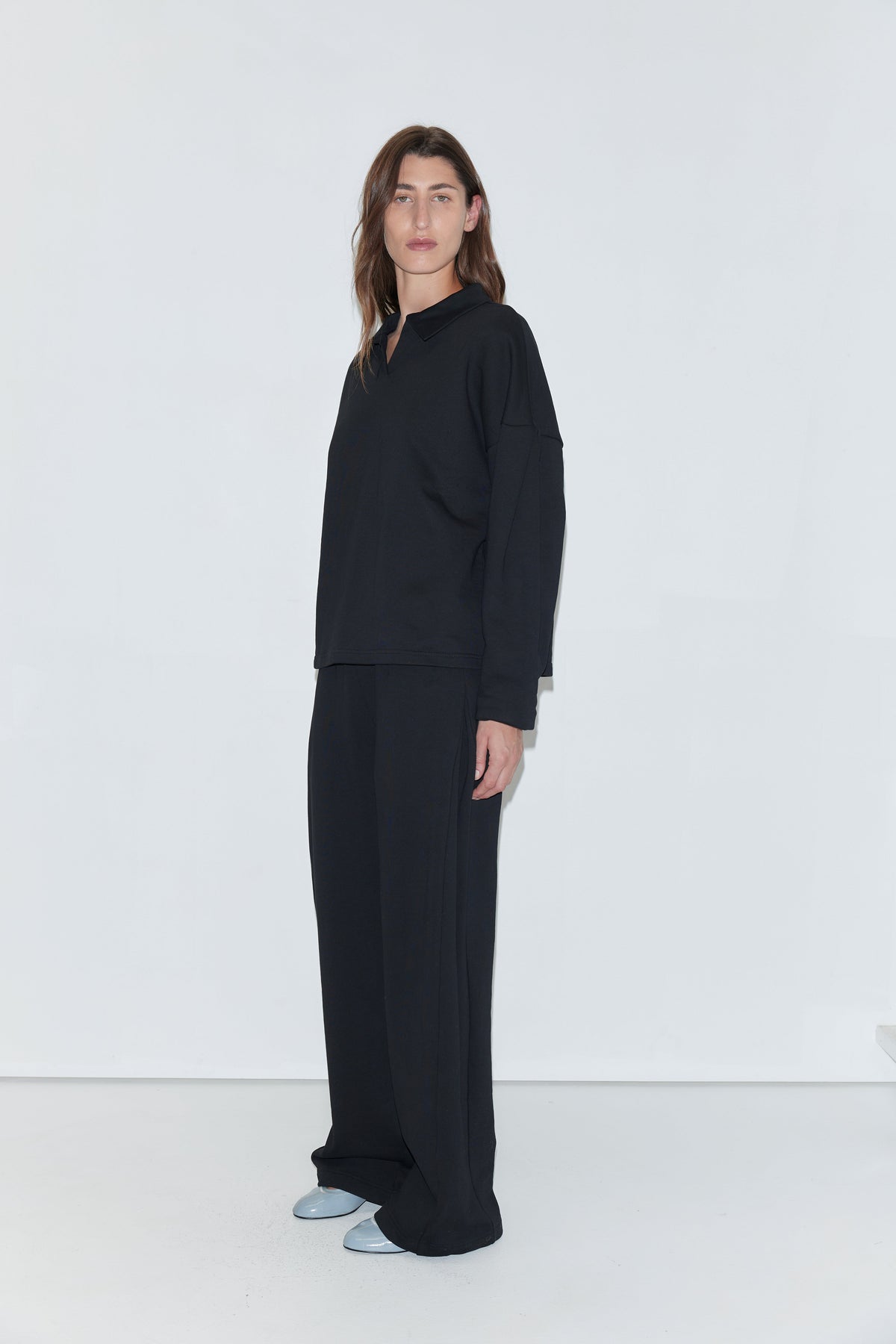 A person with long brown hair stands against a white background, wearing the Jersey Jumper in black—a loose-fitting top featuring an open polo collar—paired with wide black pants and light blue shoes.