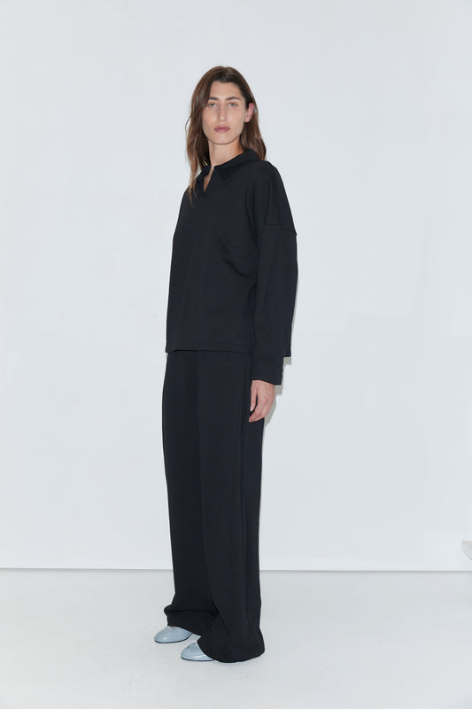 A person with long brown hair stands against a white background, wearing the Jersey Jumper in black—a loose-fitting top featuring an open polo collar—paired with wide black pants and light blue shoes.