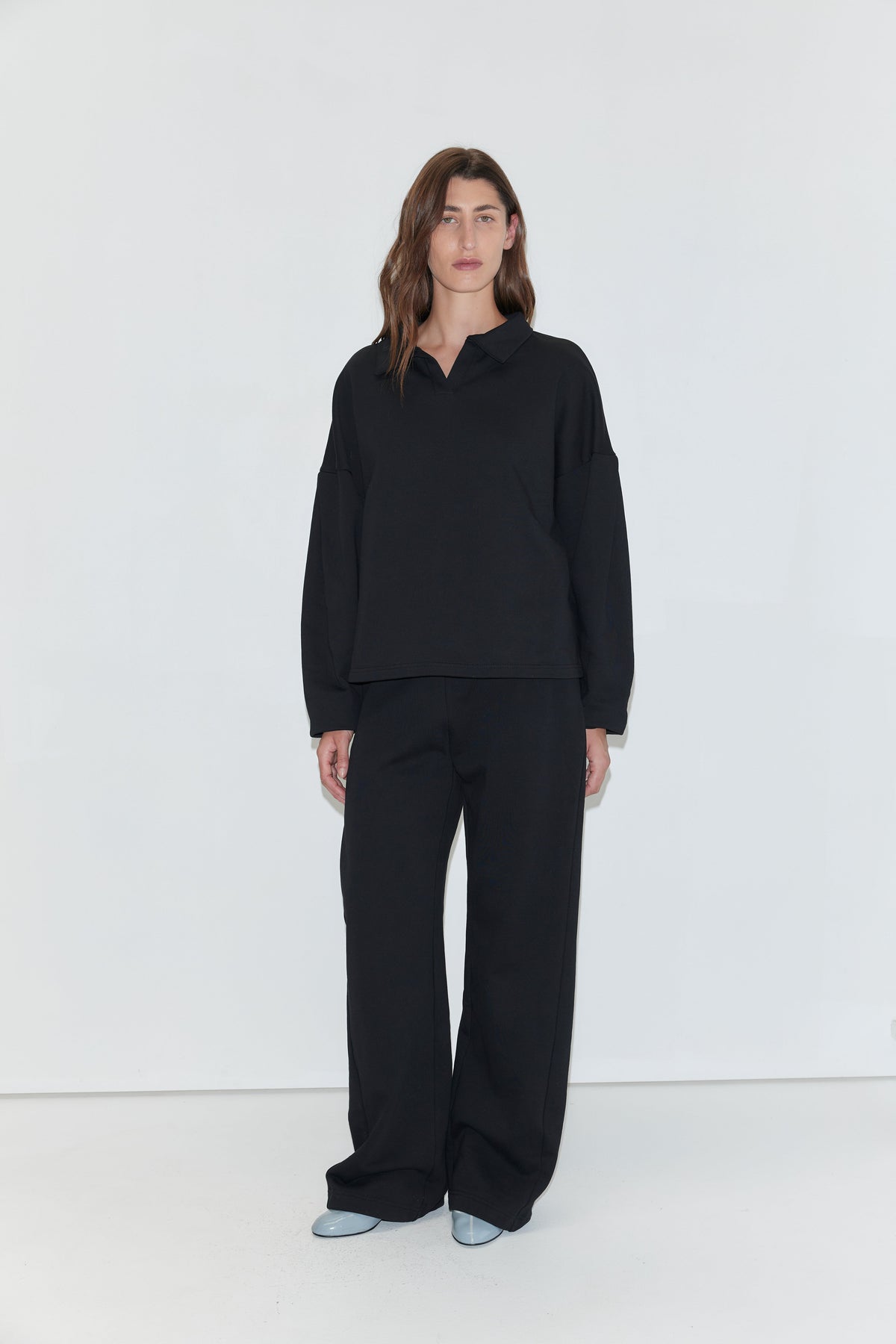 A woman with long brown hair stands facing the camera against a plain white background, wearing a black long-sleeved polo shirt and the Side Pleat Pant - Black from Deiji Studios. She is wearing light blue pointed-toe shoes.