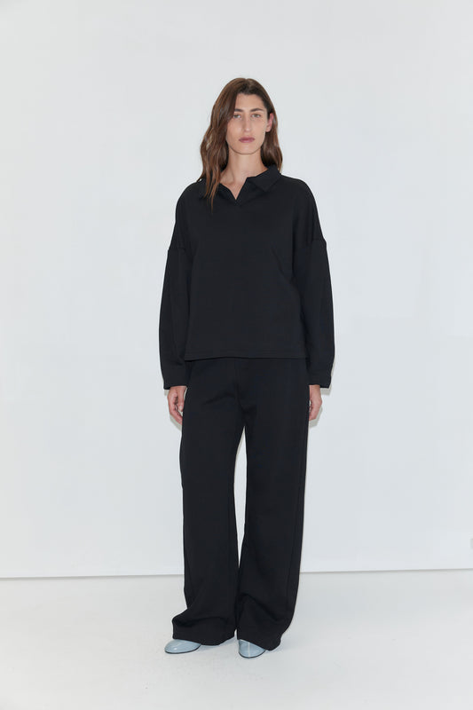 A woman with long brown hair stands facing the camera against a plain white background, wearing a black long-sleeved polo shirt and the Side Pleat Pant - Black from Deiji Studios. She is wearing light blue pointed-toe shoes.