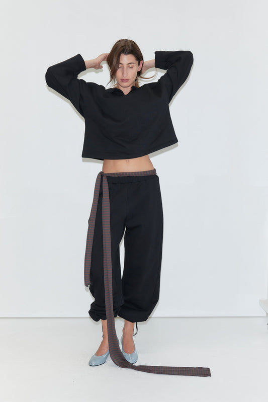 A woman stands against a white background, wearing the Jersey Jumper - Black, a loose black cotton jersey jumper with long sleeves, paired with matching black pants featuring an attached long plaid belt. She completes the look with light blue shoes and is adjusting her hair with both hands.