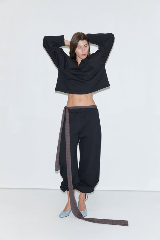 A person stands against a white background, wearing a loose black long-sleeve top, the Side Pleat Pant in black—a wide-leg cotton style with a long plaid belt reminiscent of Deiji Studios' relaxed designs—and light blue shoes, with arms raised and hands touching their head.