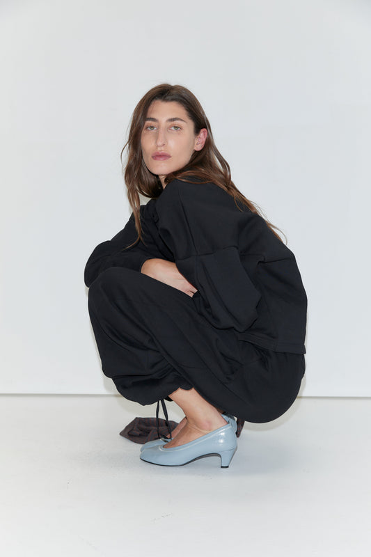 A woman with long brown hair squats against a white background, wearing the Jersey Jumper - Black and light blue heels.