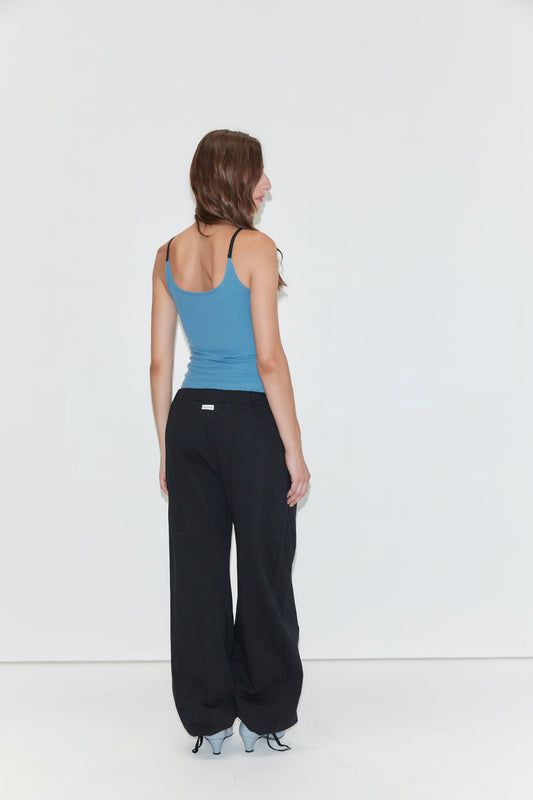 A person with long brown hair, wearing a blue tank top, Side Pleat Pant - Black by Deiji Studios, and gray shoes, stands facing away toward a white wall in a minimalist setting.