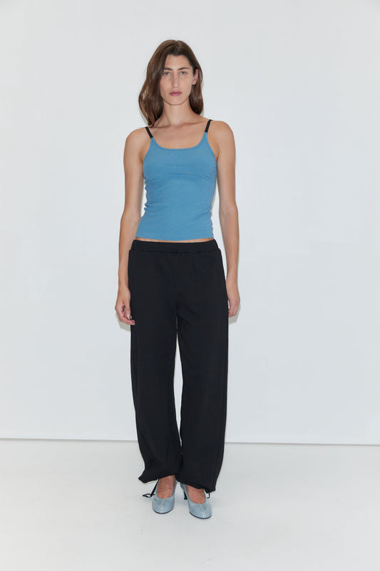 A woman with long brown hair wears a blue tank top, paired with the Deiji Studios Side Pleat Pant in black and light blue shoes. She stands against a plain white background, looking directly at the camera with a neutral expression.