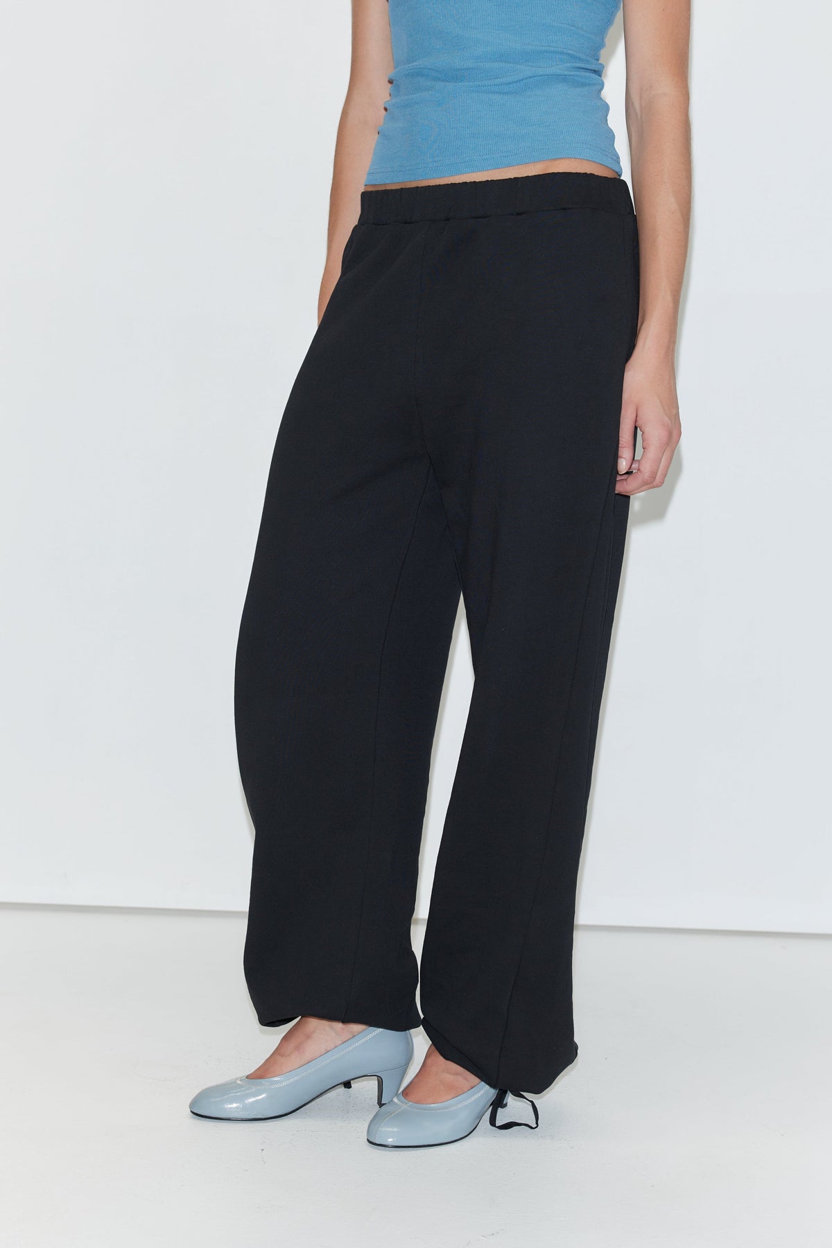 A person wearing a blue sleeveless top, Deiji Studios Side Pleat Pant - Black with an elastic drawcord, and light blue heels stands against a white background. Only the person’s torso and legs are visible.