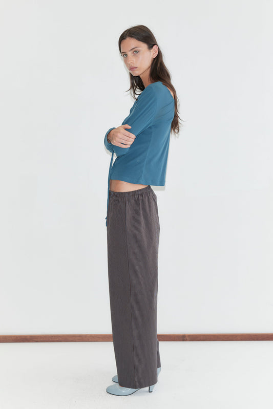 A woman with long dark hair stands in profile against a plain white background wearing the Ease Trouser - Brown Blue Stripe by Deiji Studios, dark brown wide-leg trousers with a fine blue pinstripe, with a teal long-sleeve top and pale blue heels.