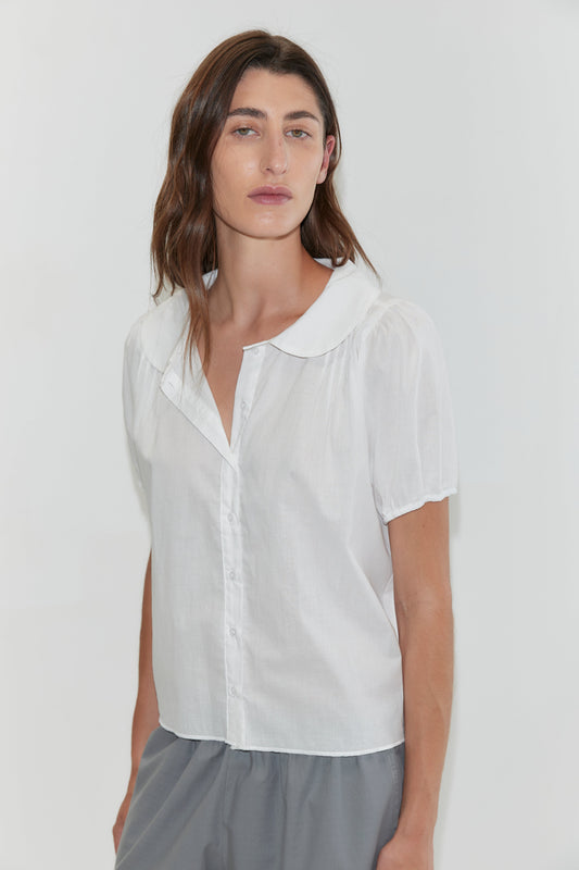 A woman with wavy brown hair wears the Petal Top in white—a short-sleeve cotton voile top with a Peter Pan collar—paired with gray pants, standing against a plain white background.