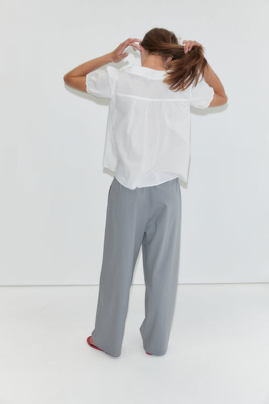 A person stands facing a white wall, holding their brown hair in a ponytail. They wear the Petal Top in White, featuring a subtle Peter Pan collar, paired with loose gray pants and red shoes. The photo is taken indoors with a minimal background, echoing the understated elegance of Deiji Studios’ style.