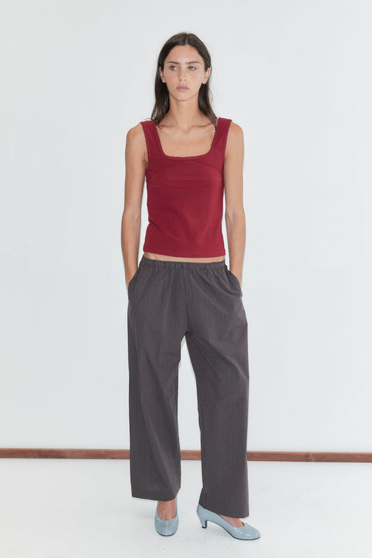 A woman with long dark hair stands against a plain white background wearing the Ease Trouser - Brown Blue Stripe by Deiji Studios, dark brown wide-leg trousers with a fine blue pinstripe, paired with a red square-neck tank and pale blue heels.